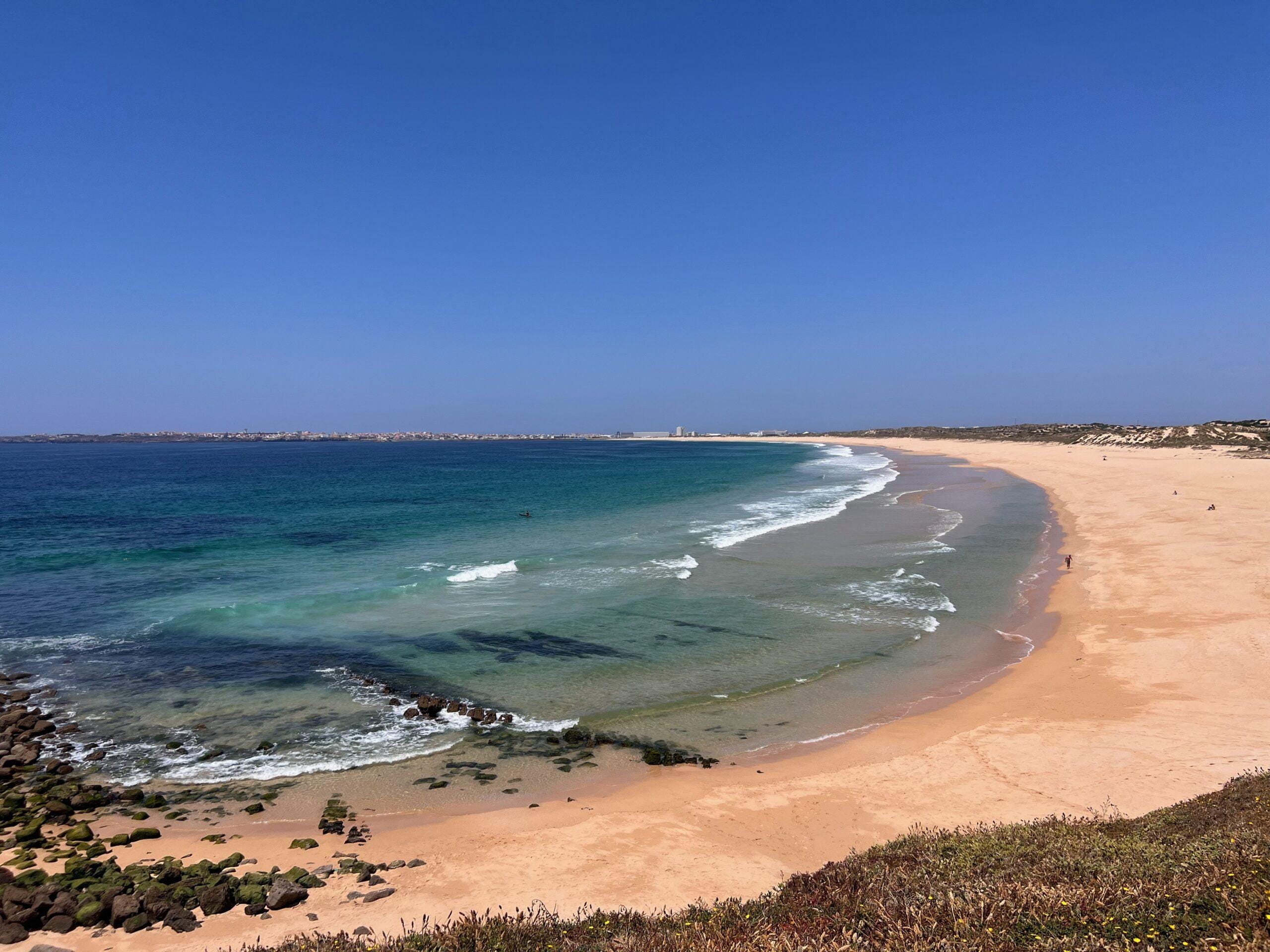 Sweeping aerial view of the wide golden sands of Praia do Consolação near Peniche, Portugal, with turquoise and deep blue Atlantic waves curving along the deserted shoreline and the town of Peniche visible in the far distance under a clear blue sky