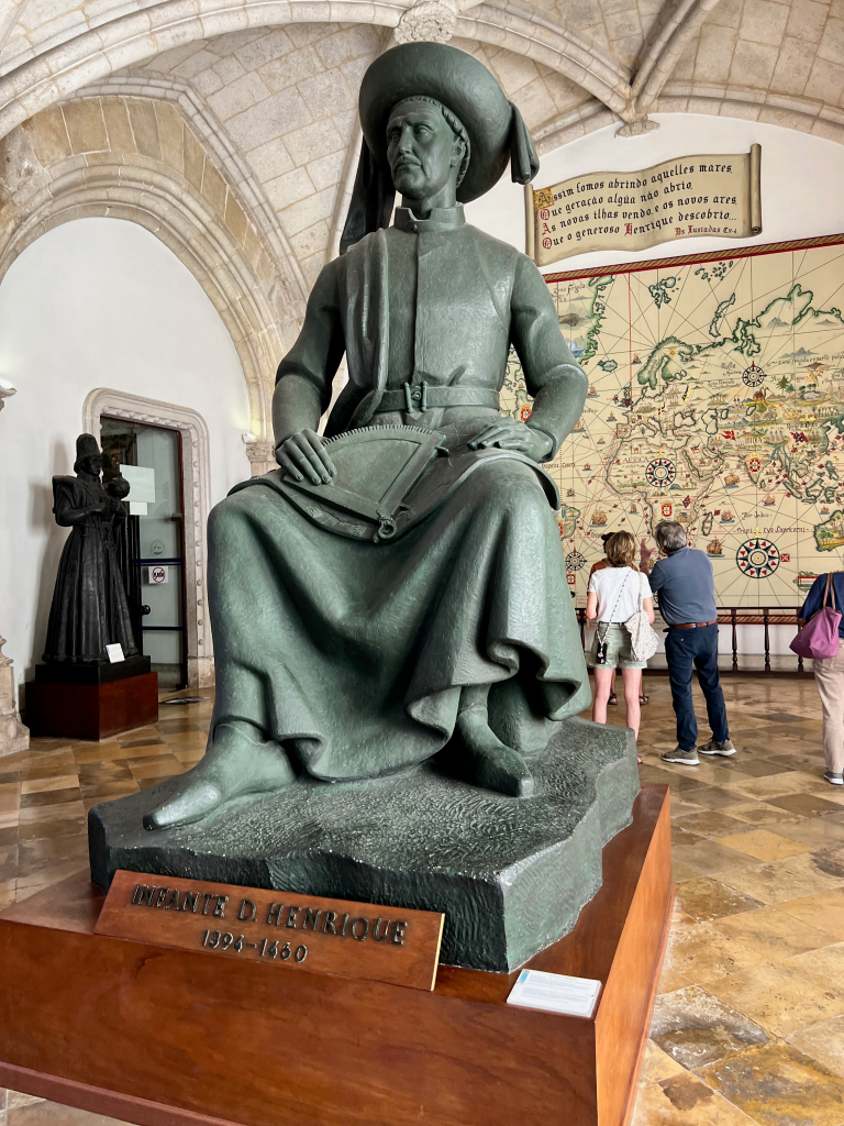 Bronze statue of Infante D. Henrique seated in a museum, wearing a wide-brimmed hat and robe, with a world map mural behind him.