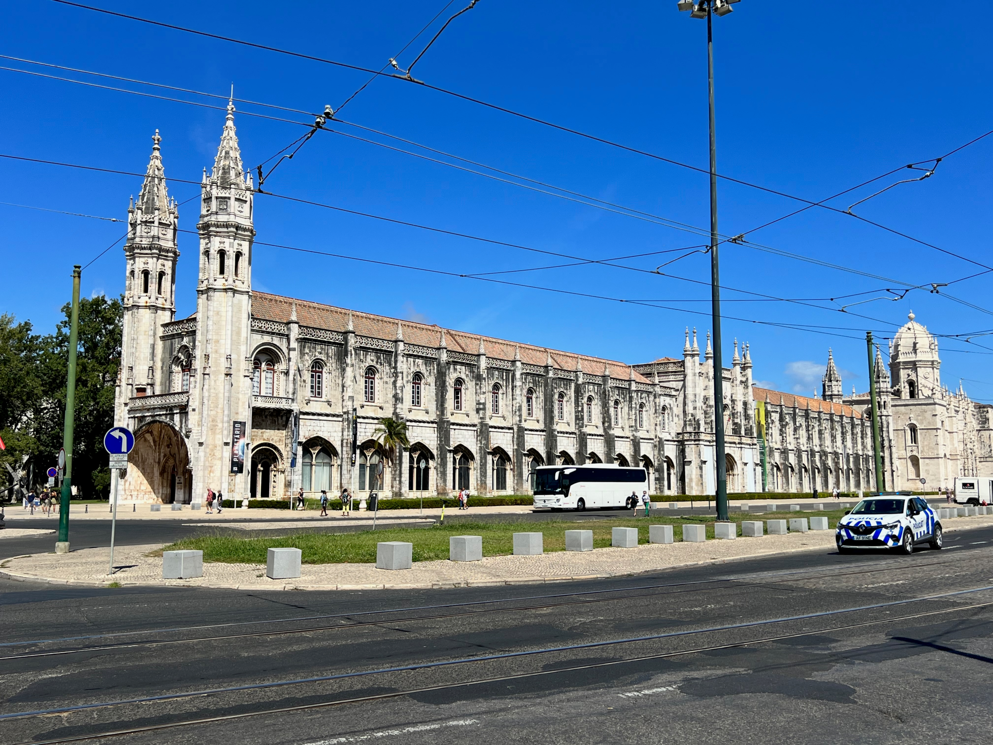 Gothic stone building with twin spires along a sunny street; overhead tram wires and pedestrians nearby.