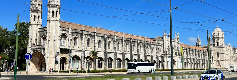 Gothic stone building with twin spires along a sunny street; overhead tram wires and pedestrians nearby.