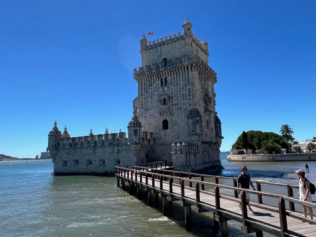 Visitors walking along the wooden boardwalk leading to the iconic Torre de Belém rising from the Tagus River in Belém, Lisbon, Portugal, with its ornate Manueline stone battlements and a Portuguese flag flying from the top — a highlight of any half-day in Belém