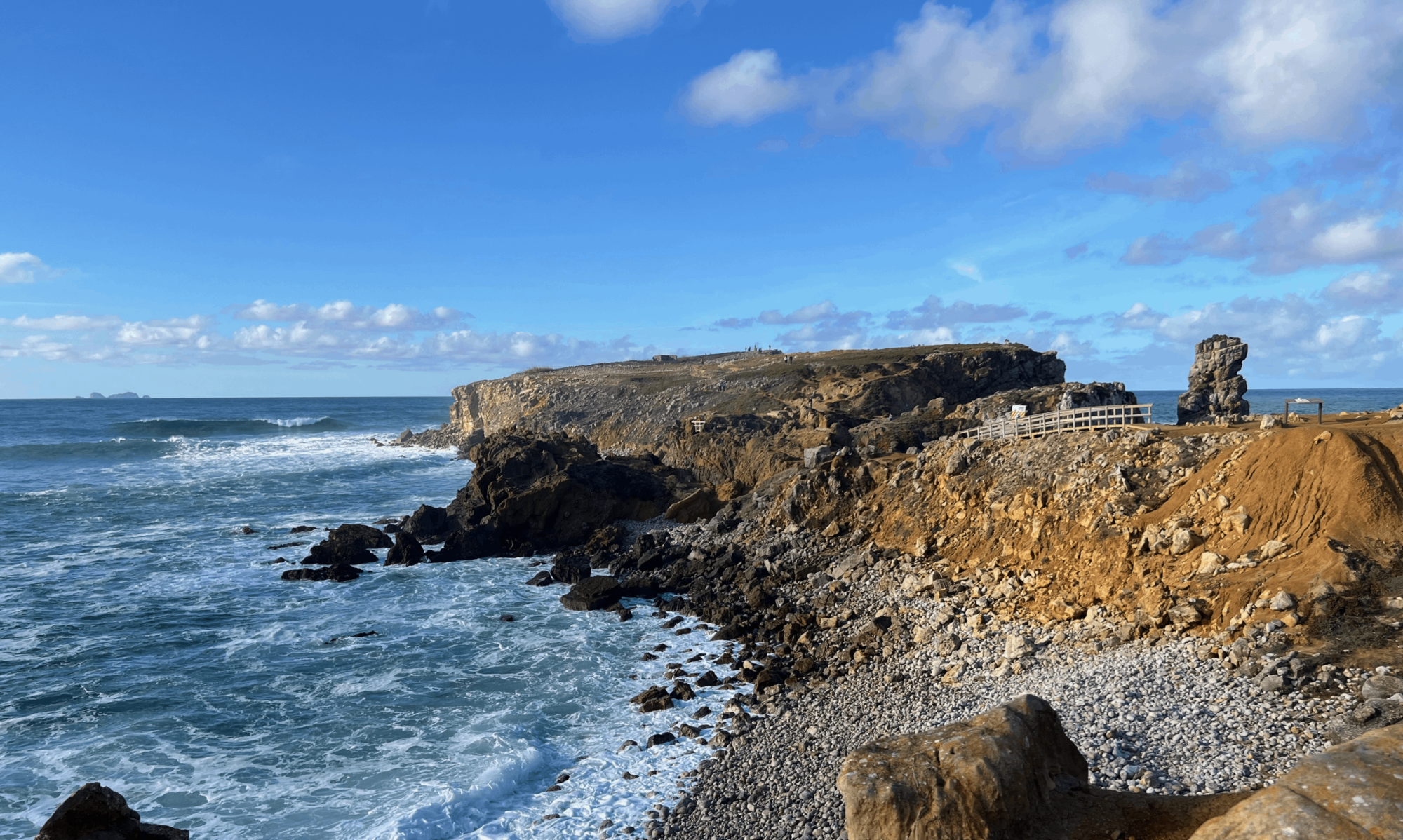 Rugged coastal cliffside with a wooden railing path along the edge and waves crashing on a pebbled shore under a blue sky with scattered clouds.